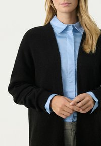 Cardigan negru tricotat, cu un design deschis în față, având mâneci lungi și o textură moale, purtat peste o cămașă cu guler de culoare albastru deschis.