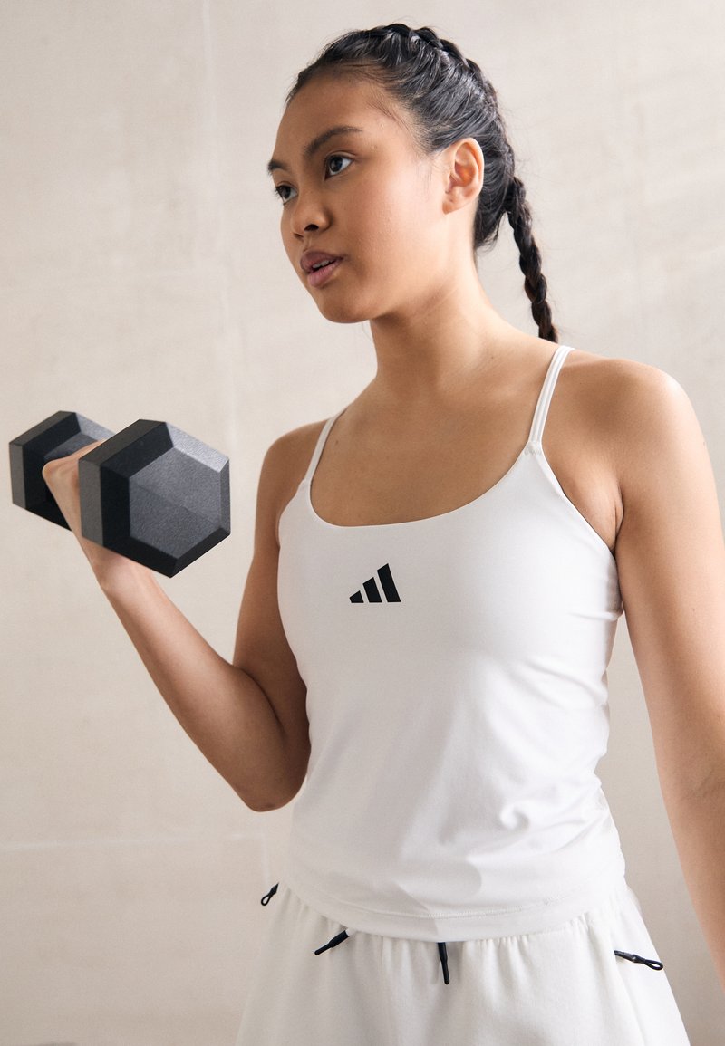 Witte sportieve tanktop met dunne bandjes en zwart logo, gecombineerd met lichtgekleurde shorts met trekkoord. Houdt een zwarte hexagonale dumbbell vast.
