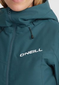 Gros plan d'une personne portant une veste O'Neill bleu sarcelle avec une fermeture éclair à l'avant et des cordons de capuche réglables visibles.