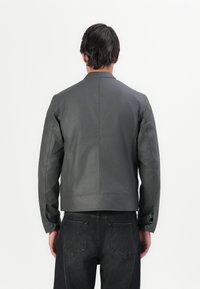 Grå lærjakke med minimalistisk design, med ståkrage og lange ermer, kombinert med svarte denim-bukser. Glatt tekstur.