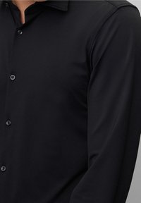 Camicia nera a maniche lunghe con bottoni, con una texture liscia, un solo bottone sul petto e un colletto classico. Nessun motivo o accento visibile.