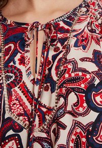 Mehrfarbige Bluse mit einem Paisleymuster in Rot und Marineblau auf einem hellen Hintergrund. Verfügt über einen Knoten-Schlüssellochausschnitt und sichtbare Nähte.
