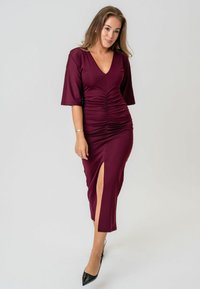 Burgundská šaty s výstrihom do V, nariasený dizajn na prednej strane, krátke rukávy a bočný rozparok; v kombinácii s čiernymi topánkami na ihlovom podpätku.