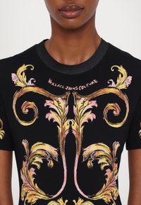 T-shirt negru cu un model floral în nuanțe de galben și roz. Decupaj rotund la gât, cu margine închisa la culoare. Textul "Versace Jeans Couture" deasupra designului.