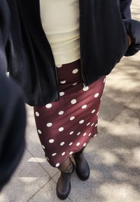 Jupe bordeaux à pois blancs, assortie à une veste noire à fermeture éclair et des bottines brunes. Tissu lisse, design ajusté.