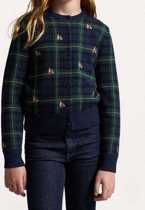 Dziecko noszące granatowy i zielony kardigan w kratę tartan z małymi brązowymi i białymi motywami psów, w połączeniu z ciemnoniebieskimi dżinsami.