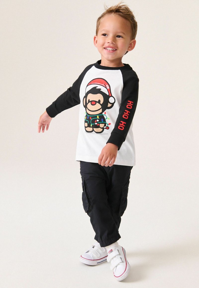 Langärmliges T-Shirt in Weiß und Schwarz mit einem Cartoon-Affen in einem Weihnachtsmannhut. Schwarze Cargo-Hosen und weiße Sneaker mit Klettverschlüssen.