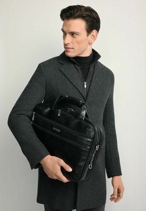 Czarna skórzana aktówka z dwoma górnymi uchwytami, widocznym zamkiem błyskawicznym i przednią kieszenią. Noszona przez mężczyznę w szarym wełnianym płaszczu, na neutralnym tle.