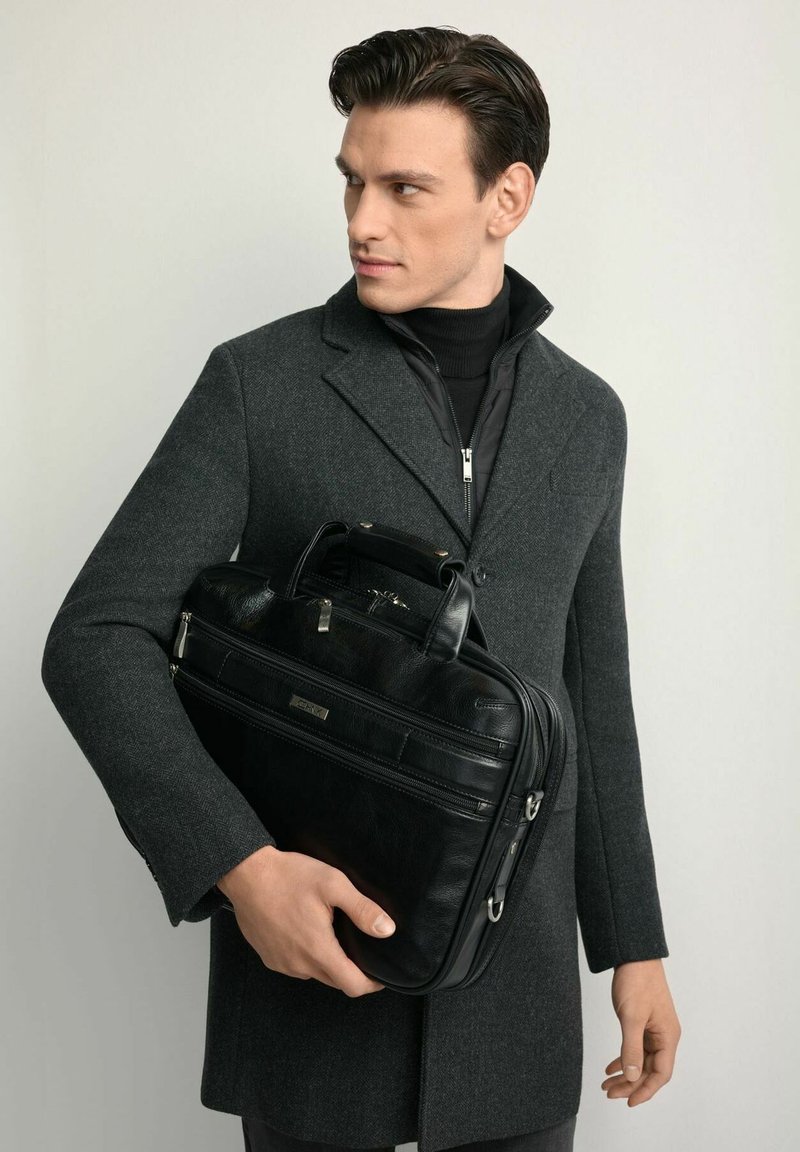 Czarna skórzana aktówka z dwoma górnymi uchwytami, widocznym zamkiem błyskawicznym i przednią kieszenią. Noszona przez mężczyznę w szarym wełnianym płaszczu, na neutralnym tle.