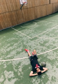 En person ligger på en grön basketplan, iklädd en svart t-shirt med en röd grafik, svarta shorts och röda sneakers. En basketboll ligger i närheten.