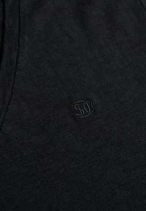 Tela negra de algodón con textura suave, que presenta un logo bordado sutil. El diseño es simple y ajustado, con cuello redondo.