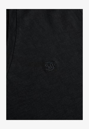 Tissu en coton noir à texture lisse, présentant un logo brodé discret. Le design est simple et ajusté, avec un col rond.