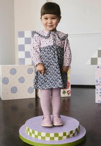 Usmievajúca sa dievča s krátkymi vlasmi v kvetinových šatách a ružových pančuchách stojí na kruhovom podstavci medzi farebnými geometrickými blokmi.
