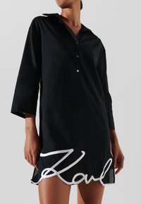 Abito camicia nero con colletto, maniche lunghe e chiusura con bottoni. Presenta testo ricamato bianco all'orlo. Tessuto leggero.
