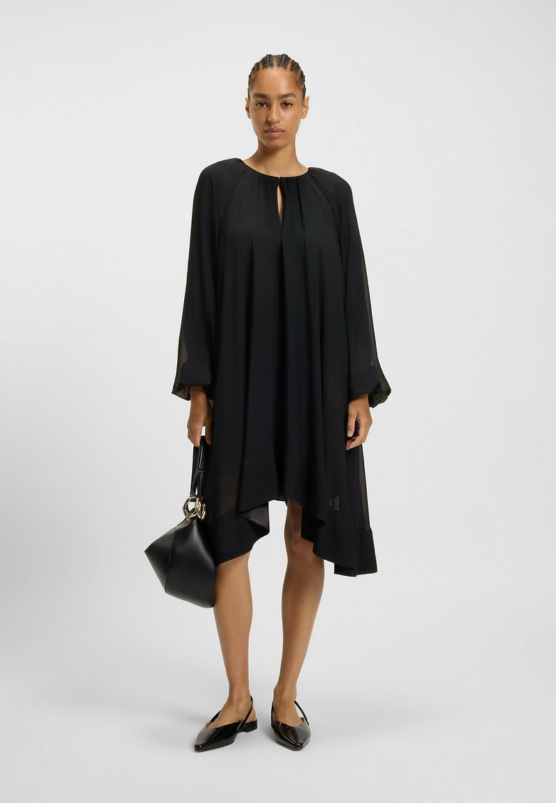 Svart, flödande klänning med långa ärmar, nyckelhålskrage och asymmetrisk nederkant. Kombinerad med en svart strukturerad handväska och spetsiga ballerinas.