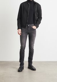 Chaqueta de denim negra con cierre de cremallera, combinada con un cuello de tortuga negro y jeans oscuros. El calzado presenta mocasines negros con costuras decorativas.