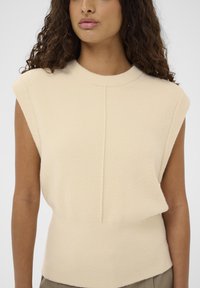 Frau trägt ein ärmelloses beige gestricktes Oberteil mit Rundhalsausschnitt und einer zentralen vertikalen Nahtdetails auf der Vorderseite.