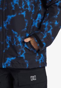 Svart jacka med blå blixtmönster, med kardborrejustering vid ärmslutet och en framficka. DC-logotyp synlig på det nedre fickområdet.