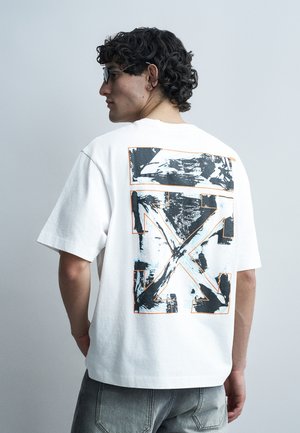 Homme aux cheveux bouclés et portant des lunettes, vêtu d'un t-shirt blanc avec un grand motif graphique abstrait noir et gris au dos.