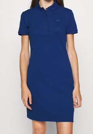 Vrouw draagt een blauw polo jurkje met korte mouwen, een knopenlijst met drie knopen en een geborduurd logo op de borst.