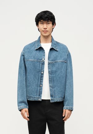 Νέος άνδρας με μπλε τζιν μπουφάν με διακριτικά γραμμικά σχέδια πάνω από άσπρο πουκάμισο και μαύρο παντελόνι, στέκεται μπροστά σε απλό φόντο.