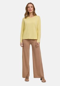 Lange mouwen gele trui gecombineerd met beige wijde broekspijpen. De trui heeft een geribbelde textuur; de broek is zacht en vloeiend. Platte sandalen maken de look compleet.