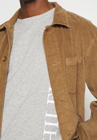 Brun kordfløjl jakke med knaplukning og brystlomme, lagd ovenpå en lys grå crewneck t-shirt med trykt tekst.