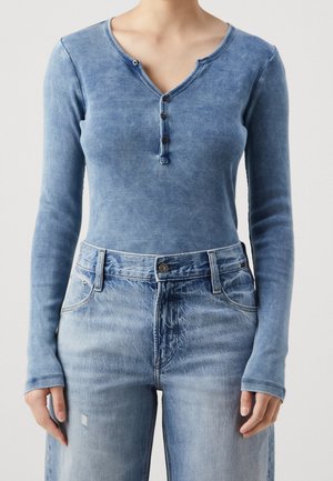 Personne portant un Henley bleu ajusté à manches longues, en maille côtelée, avec des boutons, et un jean bleu clair taille haute. Haut du corps visible.
