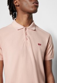 Homme portant un polo manches courtes rose clair avec trois boutons et petit logo rouge Levi's sur la poitrine gauche.