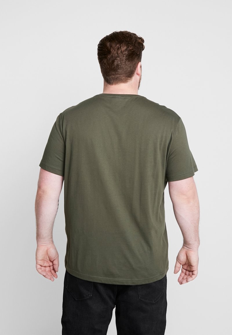 Homme de dos portant un t-shirt à manches courtes vert olive et un pantalon noir sur un fond clair uni.