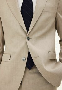 Beige kavaj med texturerad tyg, två knappar, sidofickor, slagkrage, kombinerad med en mörkblå slips med ljusa ränder.
