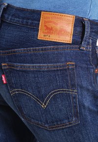 Siniset farkut, joissa on ruskea nahkainen takapäinen etiketti, jossa lukee "Levi Strauss & Co.", ja punainen tarranauha takataskussa, jossa on keltaisia ompelutietoja.