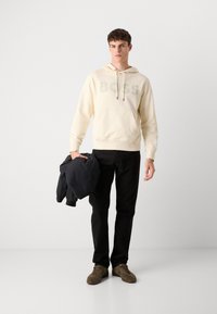 Sudadera crema con un gran logo gris de "BOSS", pantalones negros y zapatillas verde oliva. Sosteniendo una chaqueta negra con una textura suave.