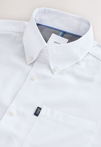 Chemise blanche à boutons avec une poche et une étiquette de marque, avec un col boutonné et une étiquette intérieure visible indiquant "NEXT."