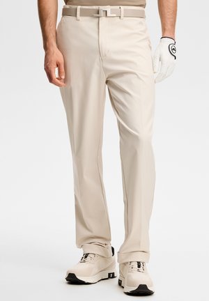 Mann trägt beigefarbene Hose, passenden Gürtel, weißen Golfhandschuh an der rechten Hand und hellfarbene Sportschuhe mit schwarzen Sohlen.