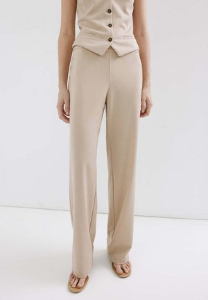 Femme portant un pantalon beige à jambes larges et un gilet boutonné assorti, avec des sandales ouvertes, debout devant un fond blanc uni.