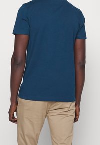 En person som bär en enkel mörkblå kortärmad skjorta och beige byxor, sedd bakifrån mot en enkel vit bakgrund.