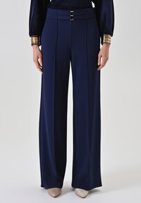 Marineblauwe high-waisted wijde pijpen broek met voorplooien, dubbele gespsluiting in de taille, gecombineerd met metallic gouden puntige schoenen en gouden armbanden.