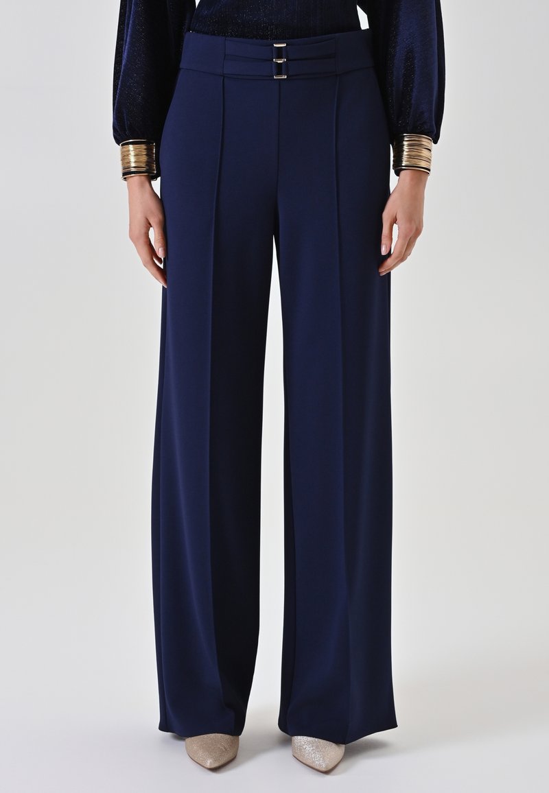 Marineblauwe high-waisted wijde pijpen broek met voorplooien, dubbele gespsluiting in de taille, gecombineerd met metallic gouden puntige schoenen en gouden armbanden.