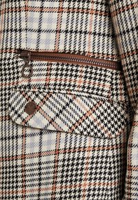 Tissu en mélange de laine avec un motif à carreaux noir, blanc et orange. Comprend une poche zippée et un accent logo métallique rond.