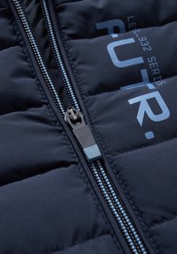 Gros plan d'un tissu matelassé bleu marine avec une fermeture éclair bleue et noire et un texte imprimé bleu partiellement visible.