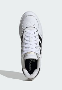 Zapatilla blanca con un upper de cuero suave, franjas negras y cordones blancos. Cuenta con una punta redondeada y un logo en la lengüeta en color dorado.