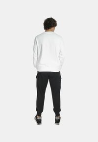 Camicia bianca a maniche lunghe, pantaloni cargo neri e sneakers nere. L'outfit presenta una vestibilità rilassata, un tessuto testurizzato e colori a contrasto.