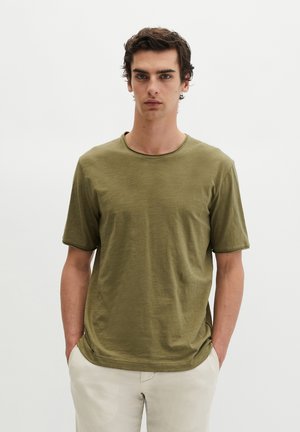 Jeune homme aux cheveux foncés portant un t-shirt vert olive à manches courtes et un pantalon beige clair, debout les mains dans les poches devant un fond uni.