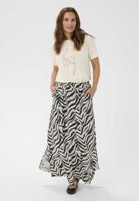 Kvinde med langt brunt hår iført en off-white blomstertrykt T-shirt og en lang nederdel med sort-og-beige zebra mønster samt sorte flade sko.