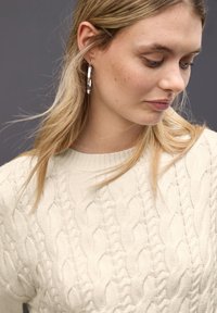 Cremfarbener Kabelstrickpullover mit rundem Ausschnitt und strukturiertem Muster. Verfügt über lange, gerade blonde Haare und einen silbernen Creolenohrring.