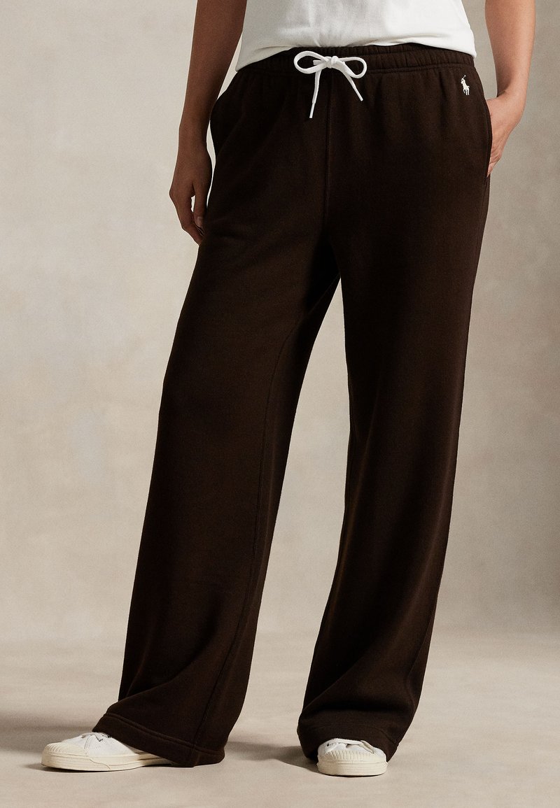 Persona che indossa pantaloni della tuta larghi marrone scuro con coulisse bianca e scarpe da ginnastica bianche, in piedi con una mano in tasca.