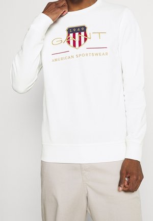 Mężczyzna noszący białą bluzę z czerwoną i niebieską tarczą oraz napisem „GANT American Sportswear 1949”, beżowe spodnie.
