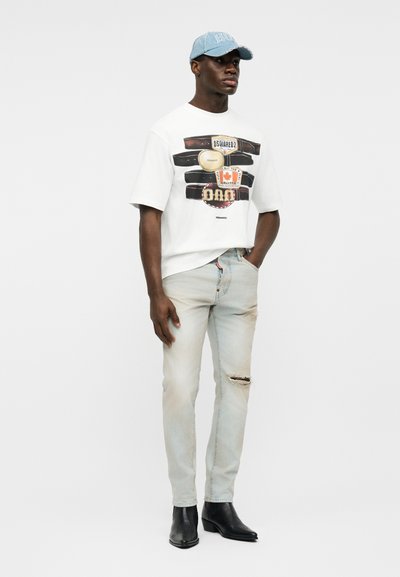 Νεαρός άνδρας που φοράει ένα λευκό γραφιστικό μπλουζάκι, ανοιχτόχρωμο σκισμένο τζιν, μαύρα μποτάκια και ένα ανοιχτό μπλε καπέλο, στέκεται με το ένα χέρι στην τσέπη.