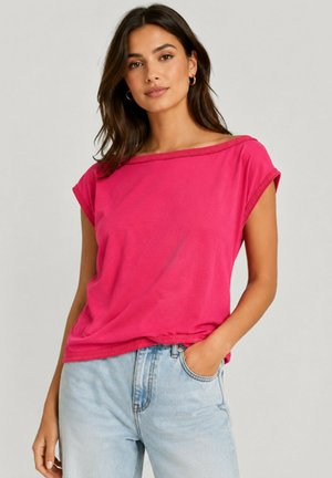 Femme aux cheveux foncés portant des boucles d'oreilles créoles dorées, un top rose vif épaules dénudées et un jean bleu clair taille haute, avec une main dans la poche.